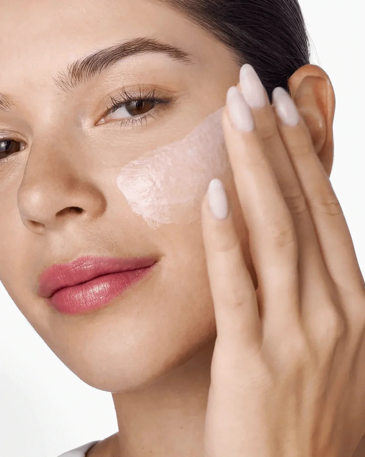 Close-up of a woman applying cream to her face with a neutral background
